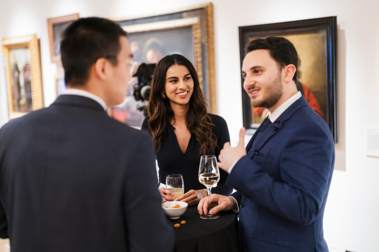 Three people in formal attire are standing around a small table at an art gallery. Two men, one giving a thumbs-up, and a woman are engaged in conversation. They have drinks and snacks on the table. Paintings hang on the walls in the background