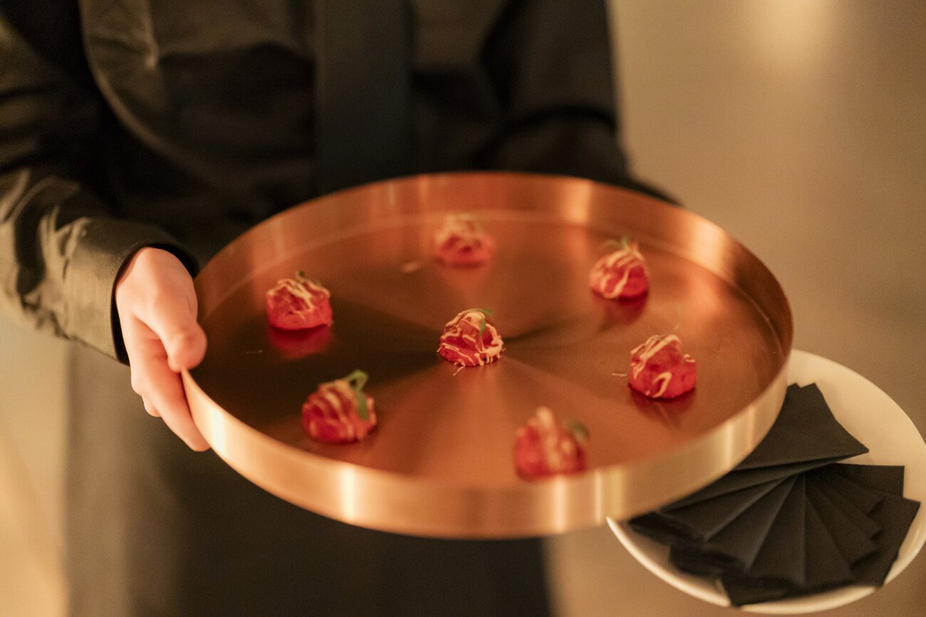 A person in a black outfit holds a copper-colored tray with eight small appetizers, possibly tomatoes or similar, topped with thin garnishes. A stack of black napkins is under the tray