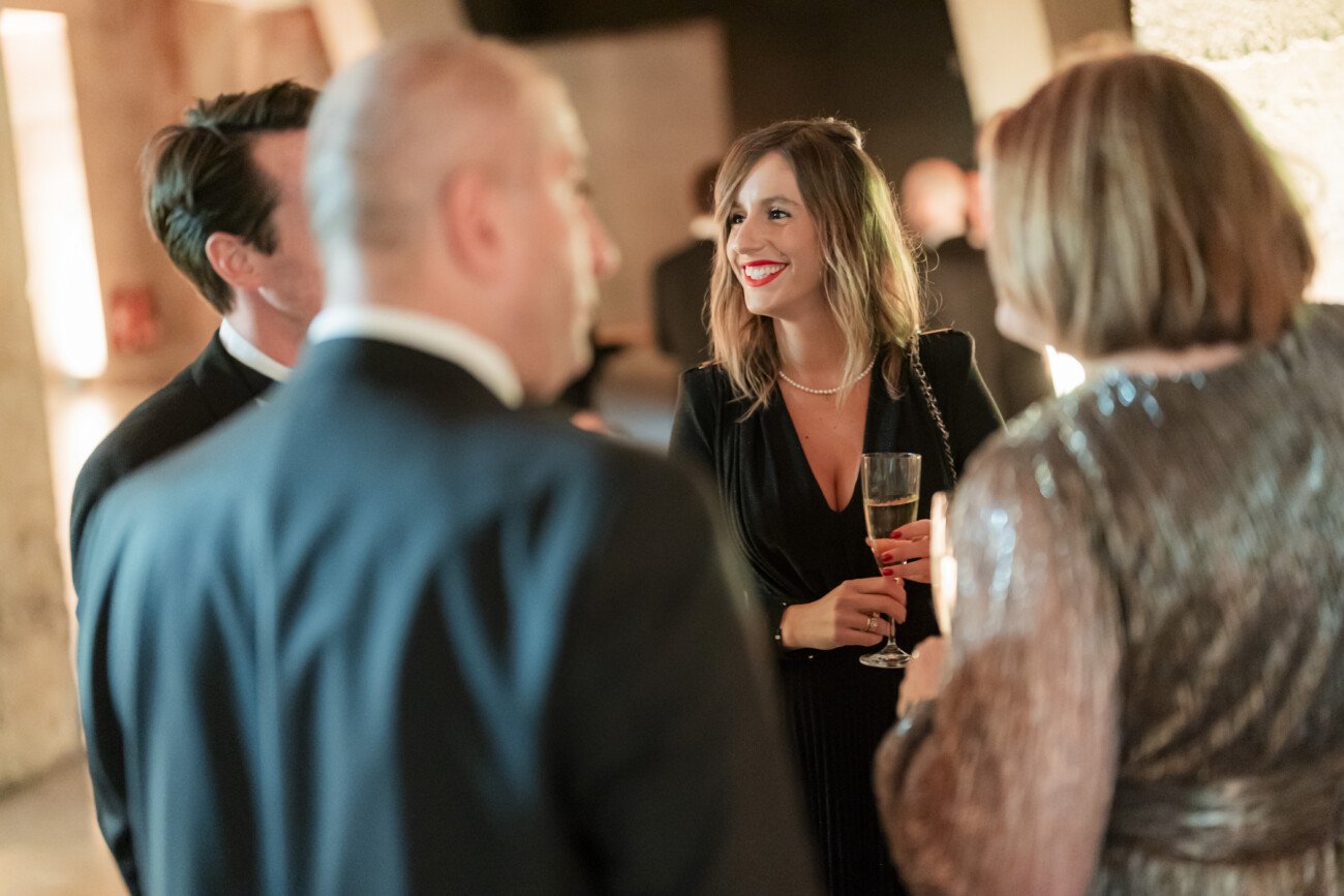 A group of people dressed in formal attire are engaged in a conversation at an event. A woman in a black dress is holding a glass and smiling. The background is softly lit, giving a warm ambiance