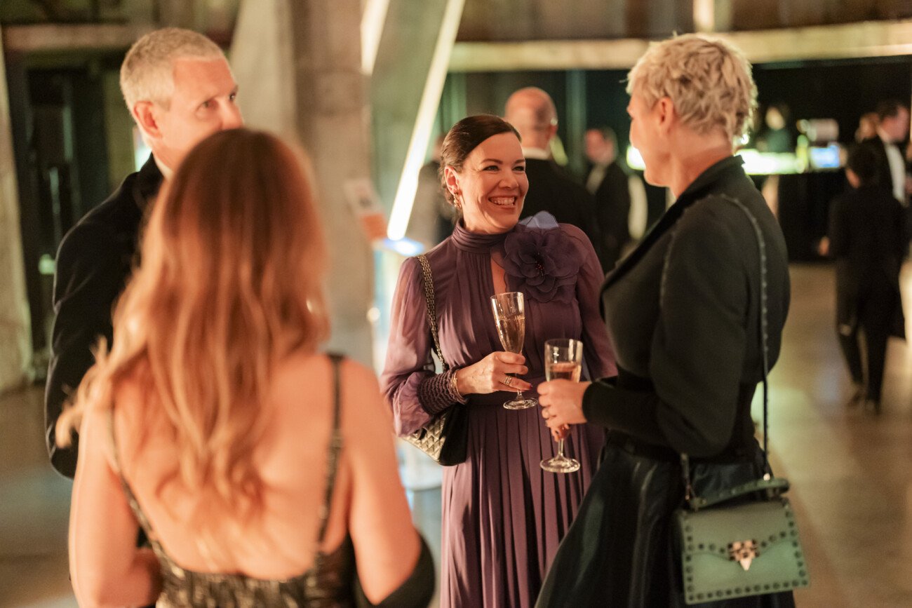 Four people dressed in formal attire are conversing at an event. Two women holding champagne glasses are smiling at each other. The background is softly lit, suggesting a formal or celebratory gathering