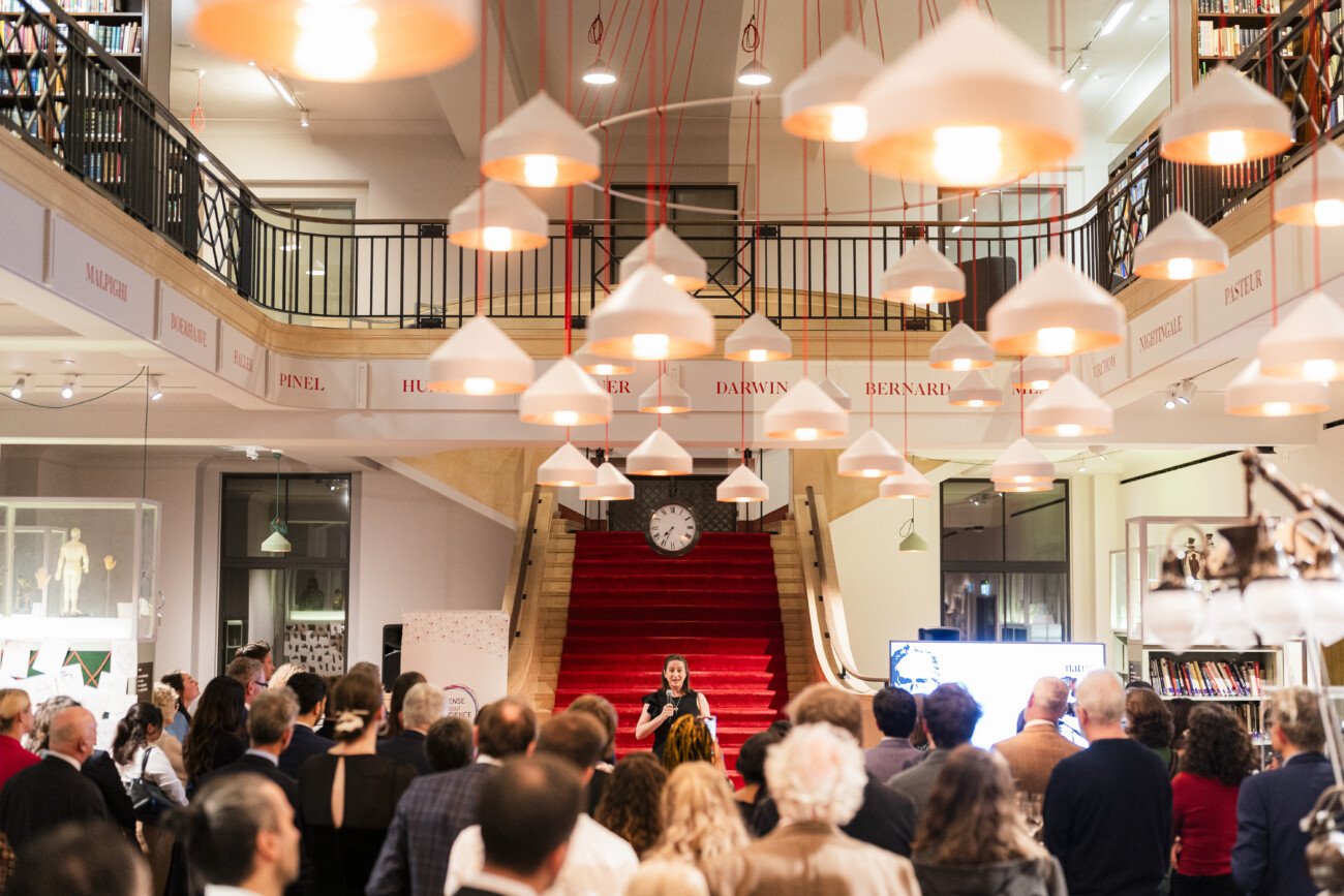 A speaker addresses a crowd in a large room with a red-carpeted staircase. Attendees face forward, surrounded by hanging lights. Shelves and displays line the walls, and a clock is visible above the staircase
