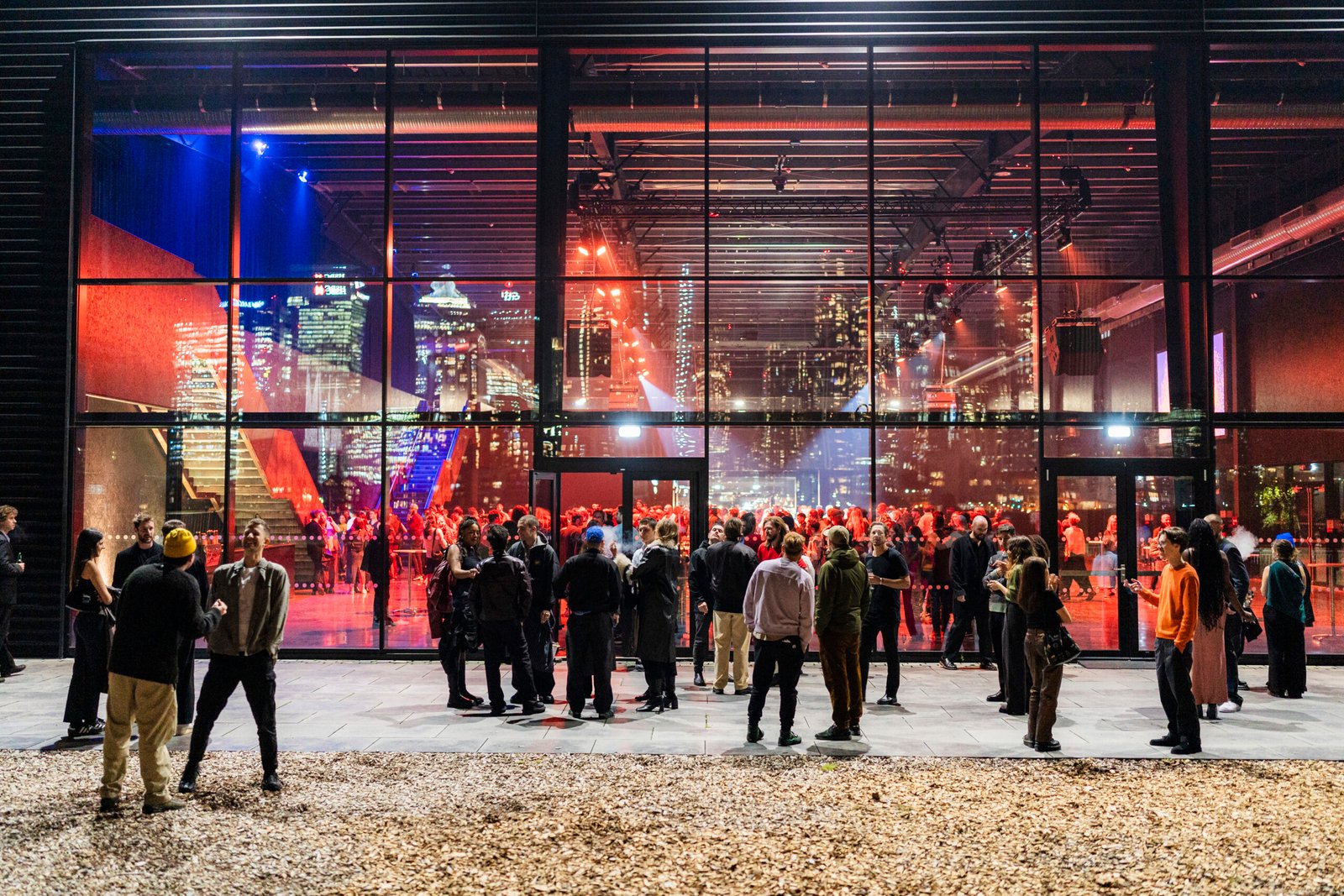 A lively crowd gathers outside a modern building with large glass windows showcasing a bustling event inside. City skyscrapers illuminate the night in the background, creating a vibrant urban atmosphere