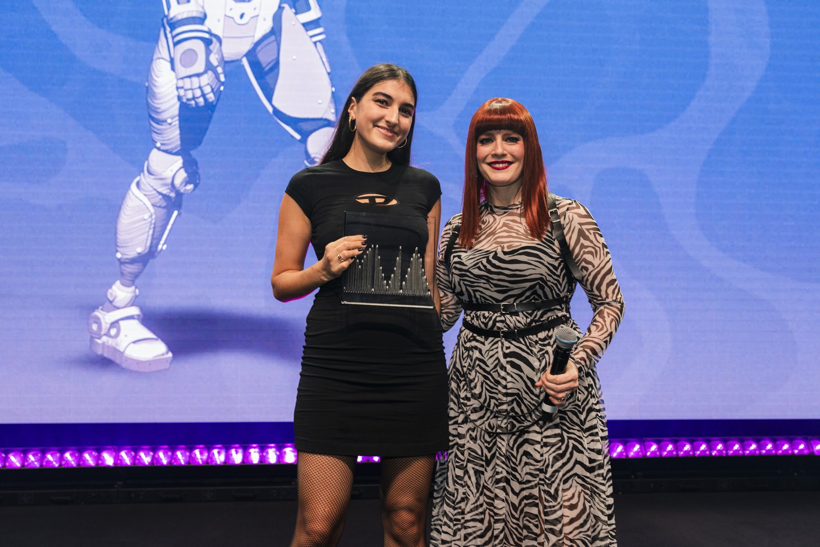 Two women stand together on stage. The woman on the left holds a trophy, while the woman on the right holds a microphone. A robot image is displayed on the screen behind them. The background is purple with abstract patterns. James Gifford-Mead Photography - Event Photographer London