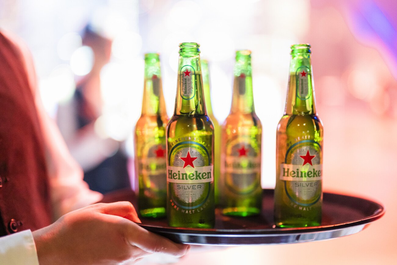 Four green Heineken Silver beer bottles on a round tray, held by a person. The background is softly blurred, highlighting the bottles