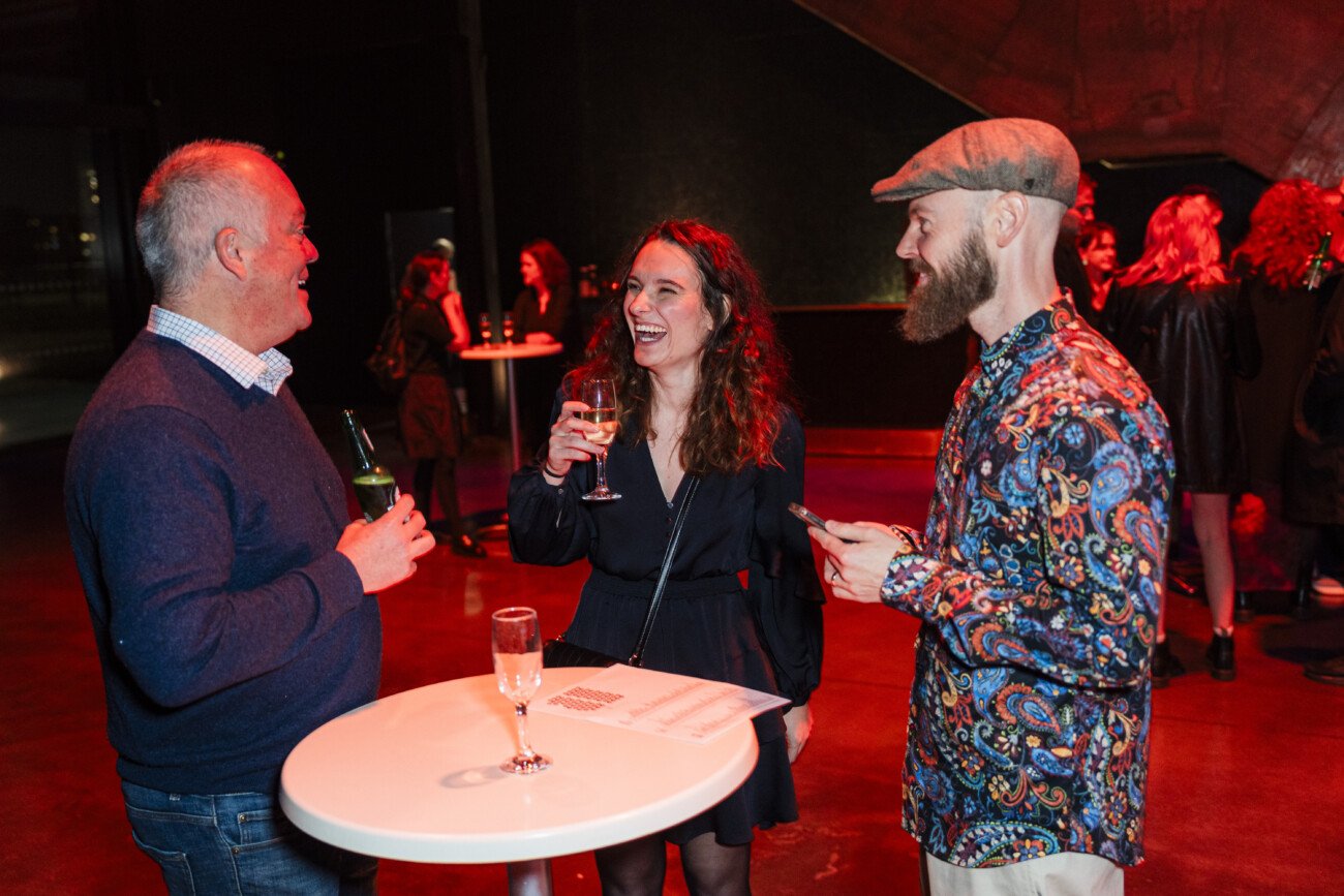 Three people are standing and laughing around a high table, each holding a drink. The setting appears to be an indoor event with a dark background and other attendees in the distance. They are dressed semi-formally