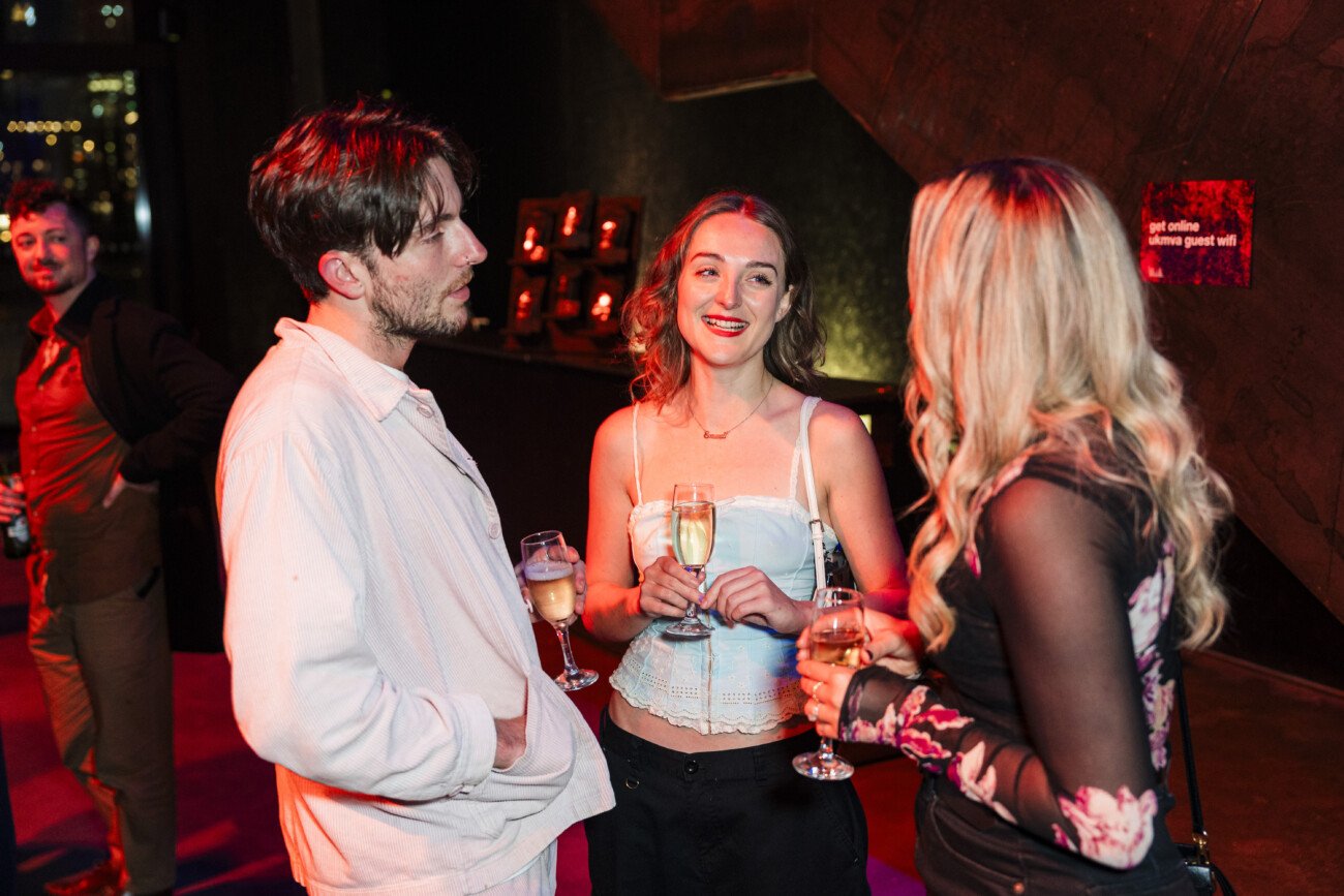 Three people are socializing at a dimly lit event, holding drinks. A man in a white shirt faces two women, one with long blonde hair in a floral top and the other with light brown hair, wearing a white strapless top. They appear engaged in conversation