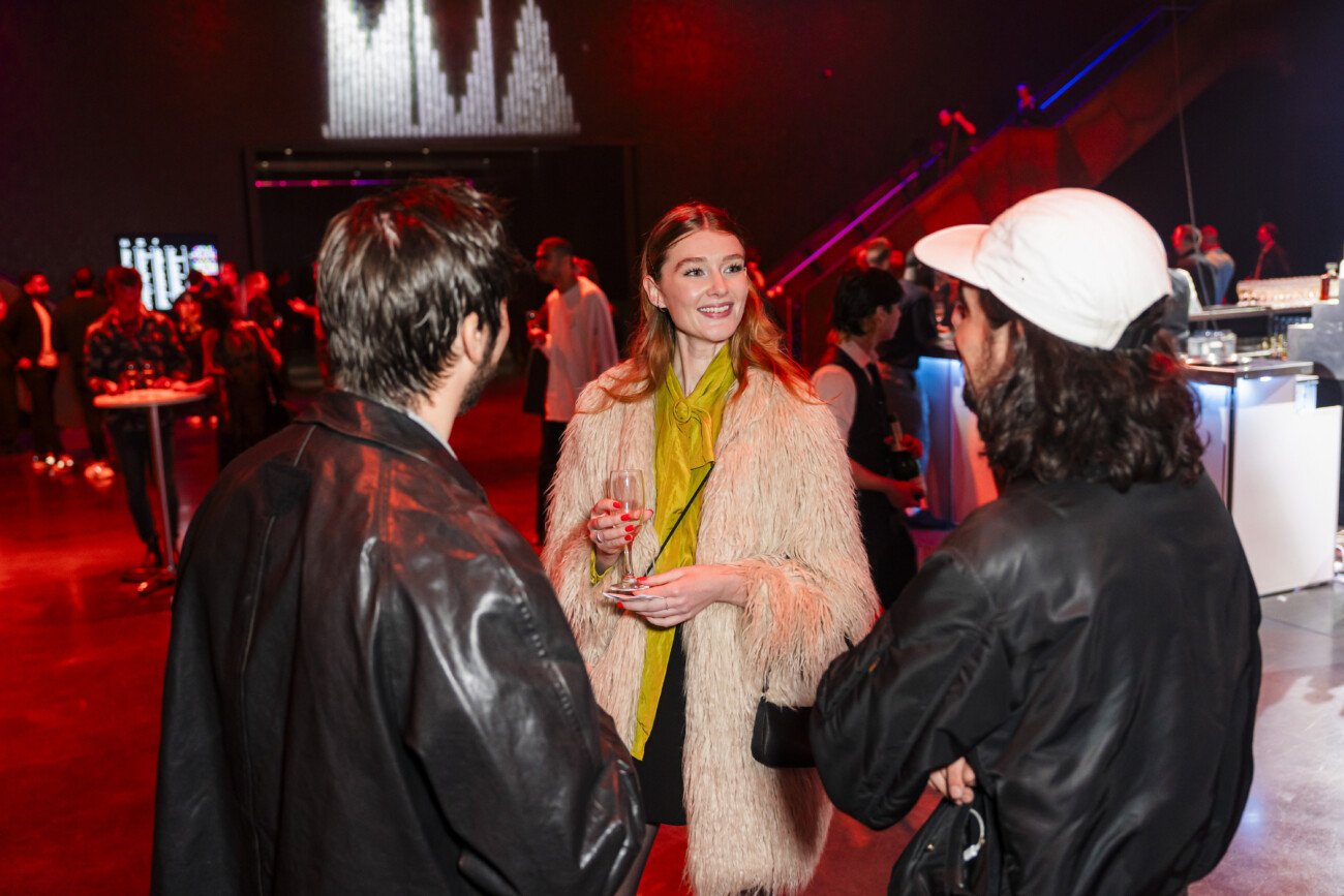 A group of three people stand in a dimly lit venue, engaged in conversation. The woman in the center wears a light-colored fur coat and holds a drink, while the two men are dressed in dark jackets. The background features red lighting and other attendees