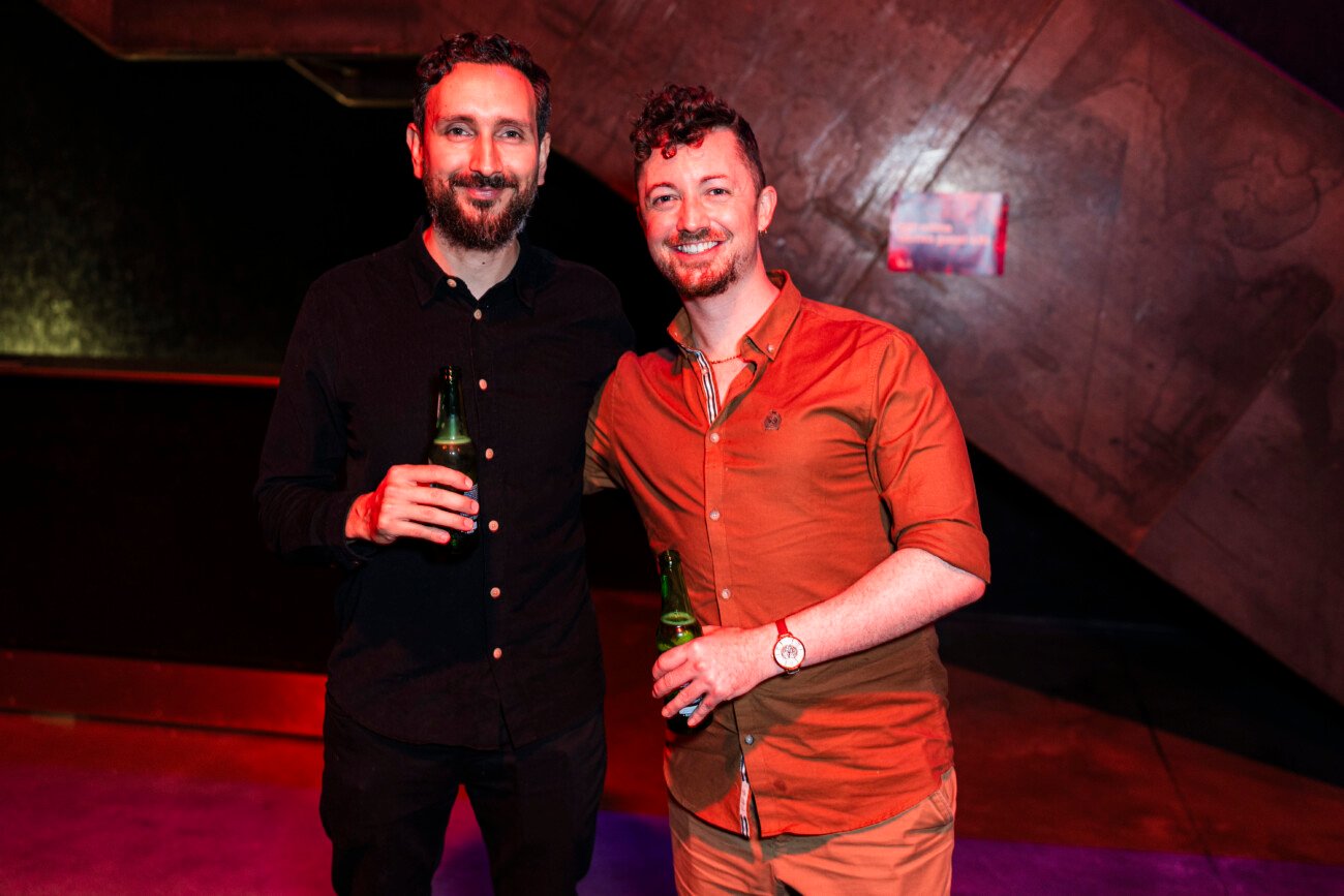 Two men smiling and holding beer bottles pose for a photo. They stand in a dimly lit room with a dark, geometric background. One wears a black shirt and the other an orange shirt