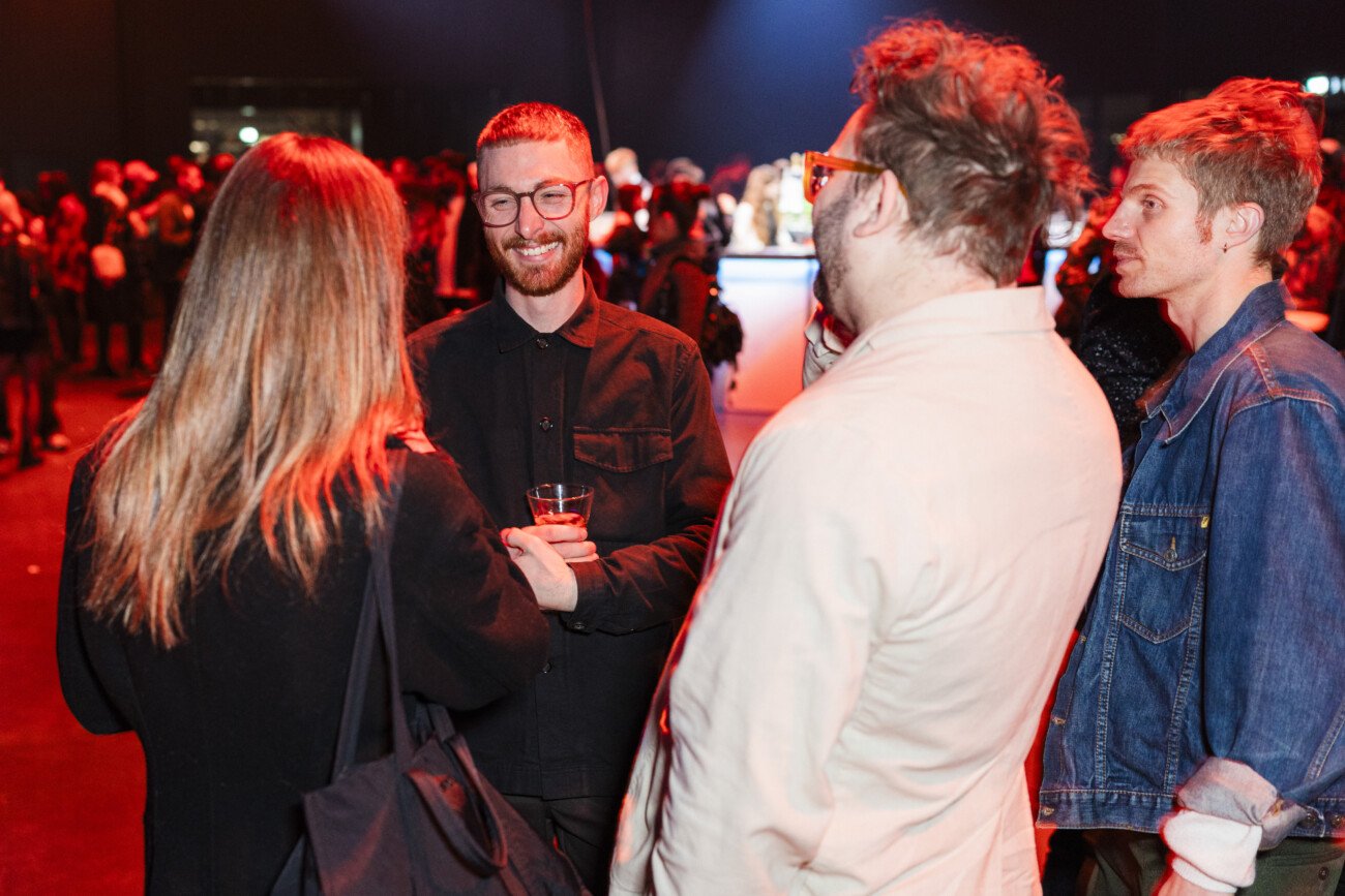 A group of people are socializing at an indoor event. One person is holding a drink and smiling, while the others listen and engage in conversation. The setting has dim lighting with a red tint, and a crowd is blurred in the background