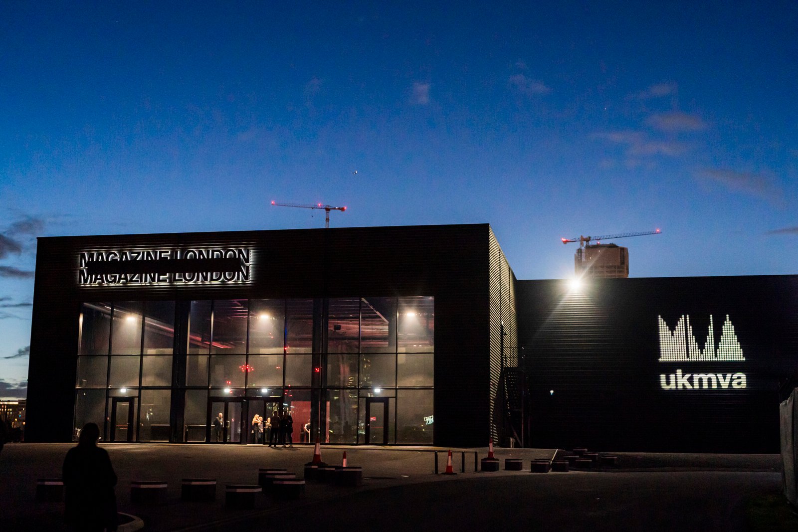 Exterior of Magazine London at dusk with illuminated signage. Two cranes are visible in the background, and the facade is lit up. The ukmva logo is projected on the building. A few people stand near the entrance