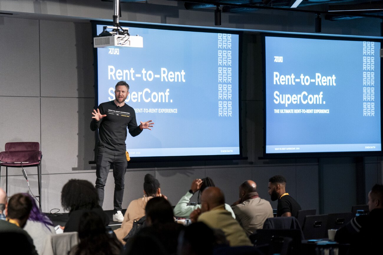 A speaker stands on stage at a conference, gesturing while addressing the audience. Behind him, two large screens display "Rent-to-Rent SuperConf." Attendees are seated, listening attentively. The environment is a modern conference room