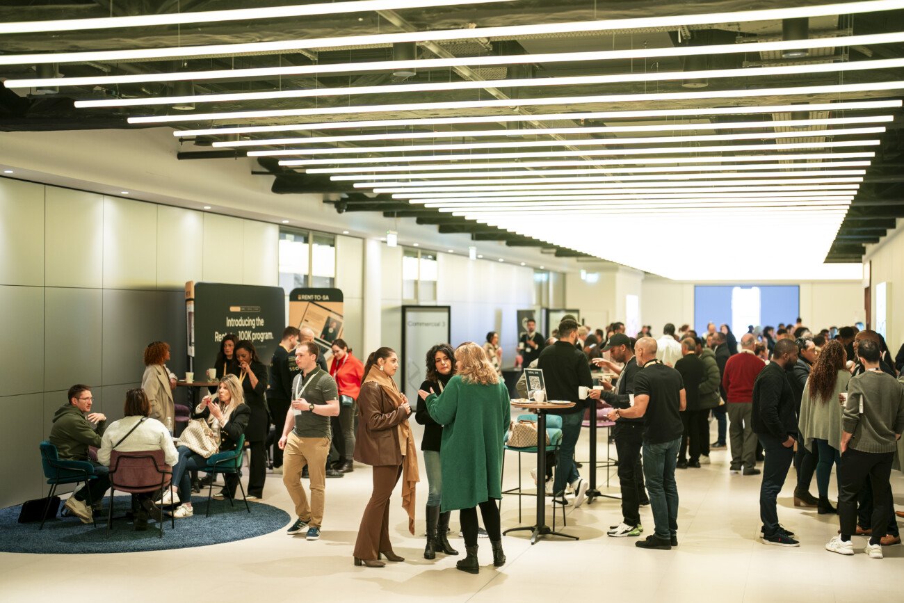 A large group of people mingling and networking in a modern, well-lit conference area. Attendees are engaging in conversations, with some seated and others standing around high tables. Overhead lights and informational banners are visible