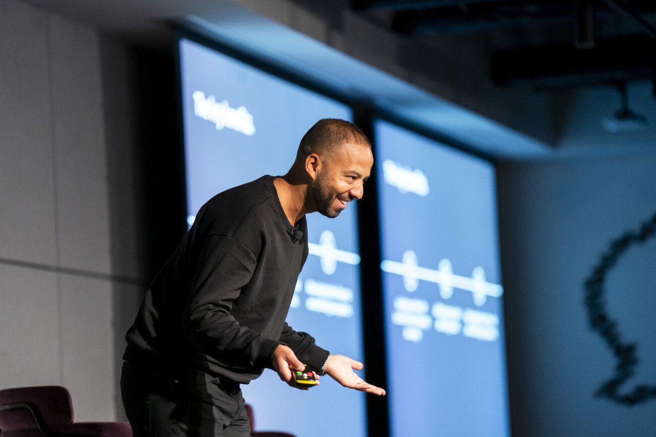 A person wearing a black sweater is speaking energetically on stage. They are holding a presentation clicker and standing in front of two screens displaying graphs and text. The setting appears to be a conference or seminar