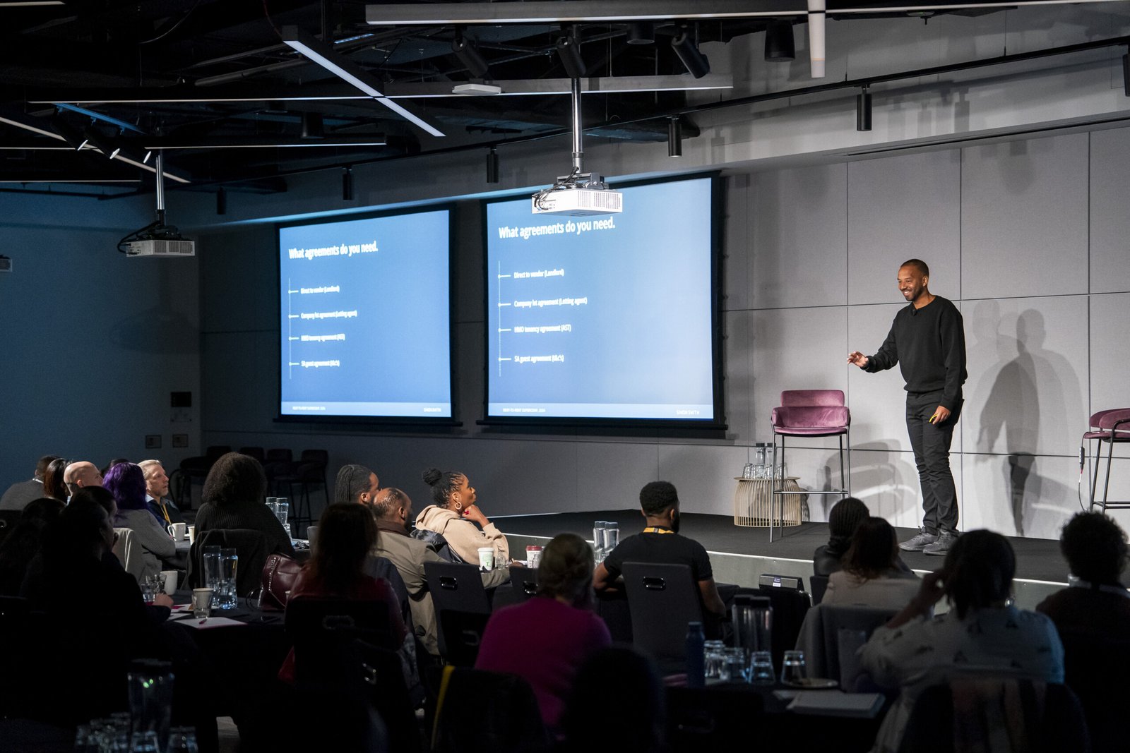 A speaker in a black outfit stands on a stage in front of an audience. Two large screens display a presentation titled "What agreements do you need." The audience is seated at tables with notebooks and glasses of water