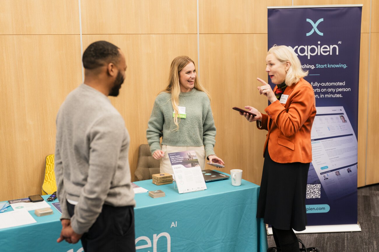 Two women stand at a booth with a blue tablecloth, engaging with a man. One woman gestures with her phone. A banner with the word "Xapien" is visible in the background. The setting appears to be a conference or event. James Gifford-Mead Photography - Event Photographer London