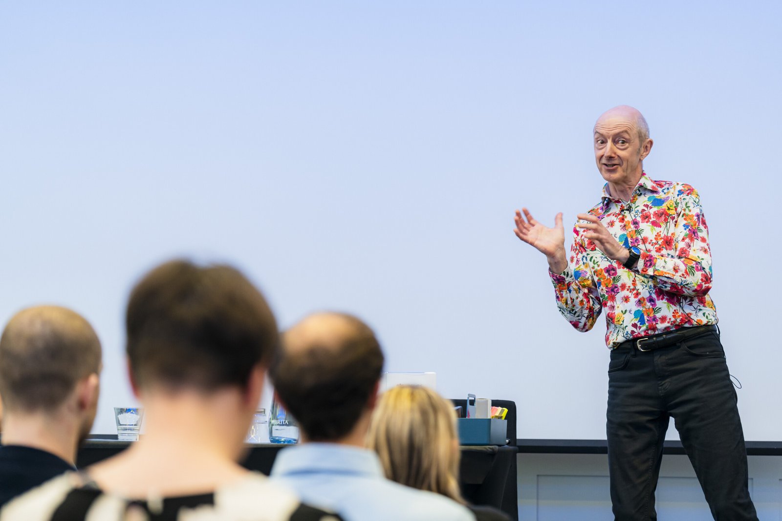 A man in a colorful shirt gestures enthusiastically while speaking to an audience. The audience's backs are visible in the foreground. He's standing in front of a white backdrop, possibly giving a presentation or lecture