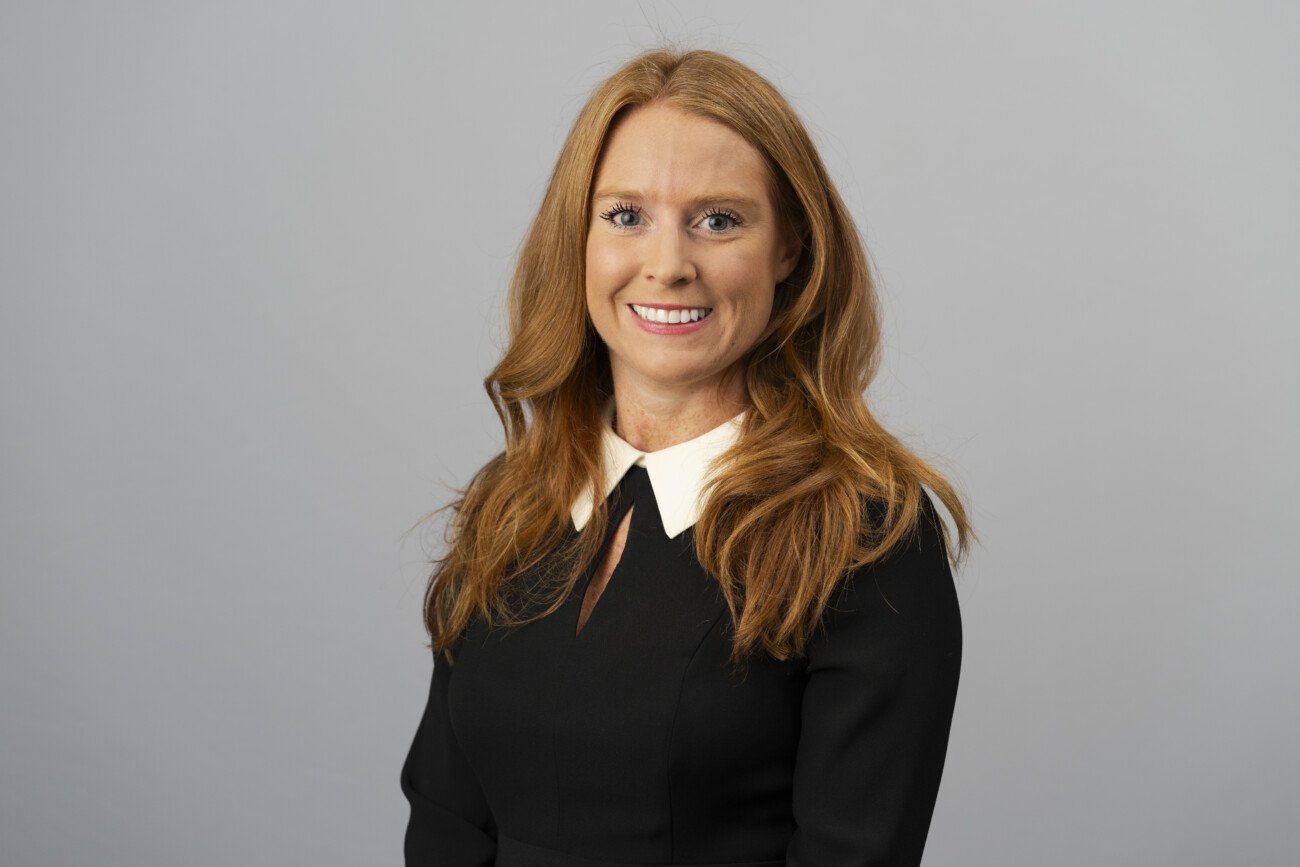 A person with long red hair smiles confidently, standing against a plain gray background, showcasing a black outfit with a white collar. This image captures the polished professionalism that a corporate headshot photographer aims to achieve. James Gifford-Mead Photography - Event Photographer London