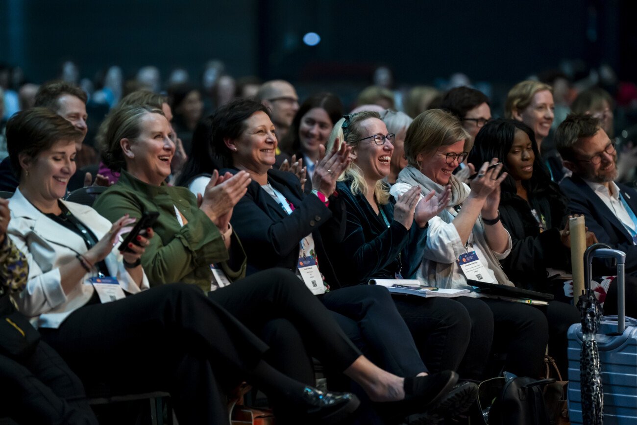 A group of people sitting in an audience, clapping and smiling. Captured through the lens of an event photographer in London, they are dressed in business attire and appear engaged and happy, seated in rows at a professional conference