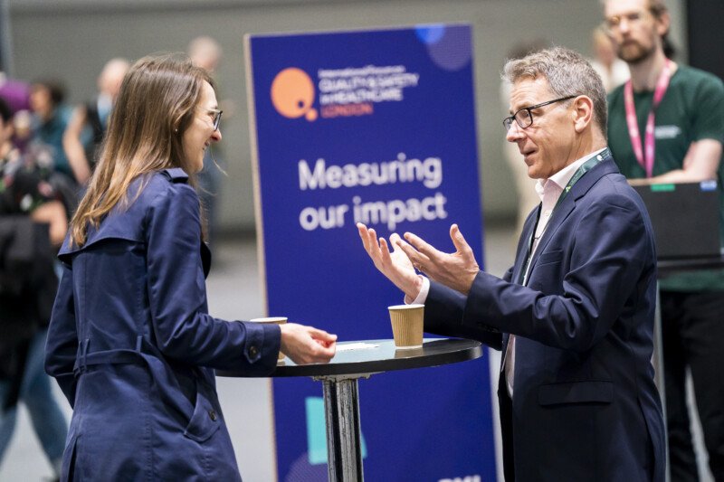 Two individuals are engrossed in conversation at a high-top table, framed perfectly by the lens of a skilled event photographer. Behind them, a sign reads "Measuring our impact," capturing the lively atmosphere of this corporate gathering. Both hold coffee cups, fully animated and engaged