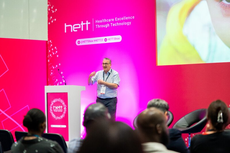 A speaker presents at a healthcare technology conference, captured brilliantly by a London event photographer. The stage, set against a pink backdrop with the "HETT" logo and tagline "Healthcare Excellence Through Technology," is abuzz as the audience eagerly listens from their seats