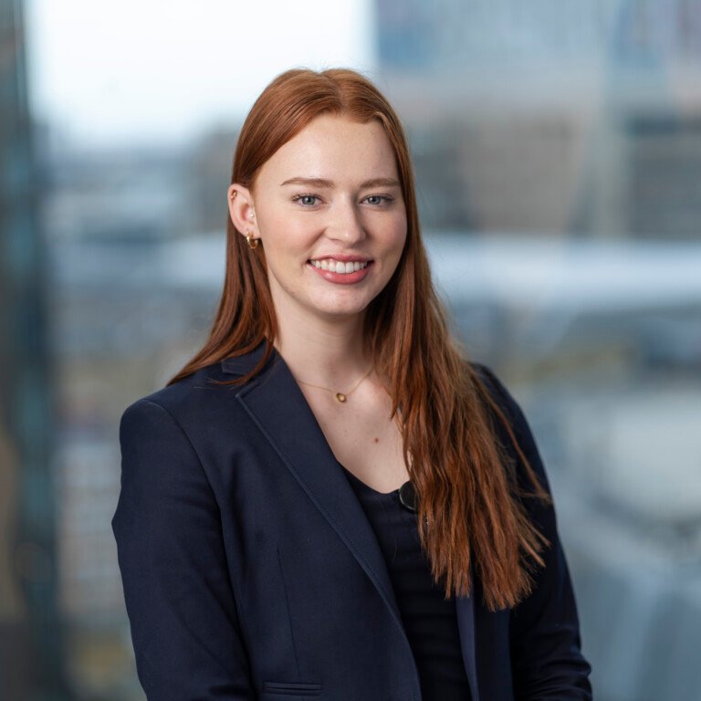 A person with long red hair is smiling, wearing a navy blazer over a black top. Captured by a corporate headshot photographer, they are indoors with a blurred cityscape visible through large windows in the background