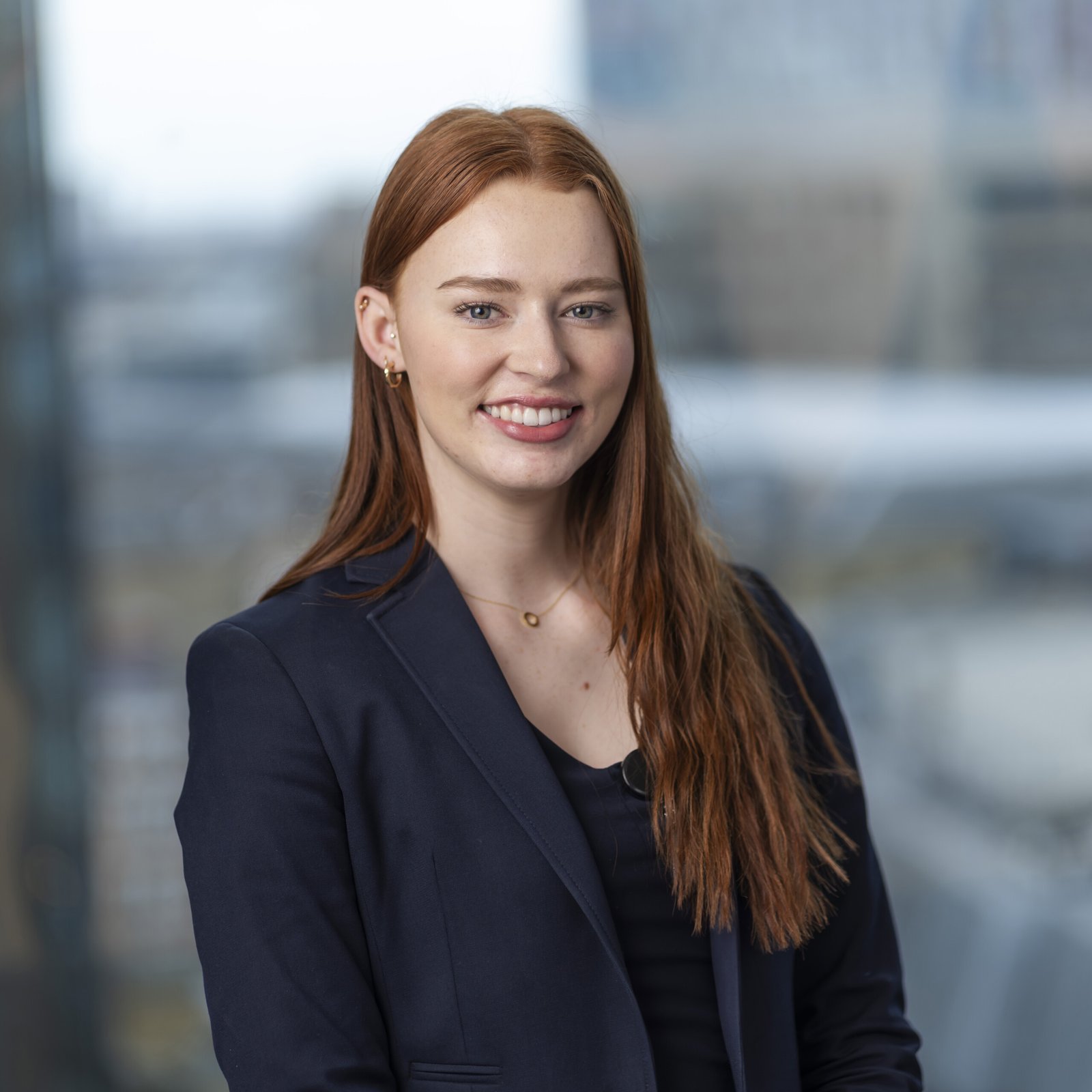 A person with long red hair is smiling, wearing a navy blazer over a black top. Captured by a corporate headshot photographer, they are indoors with a blurred cityscape visible through large windows in the background
