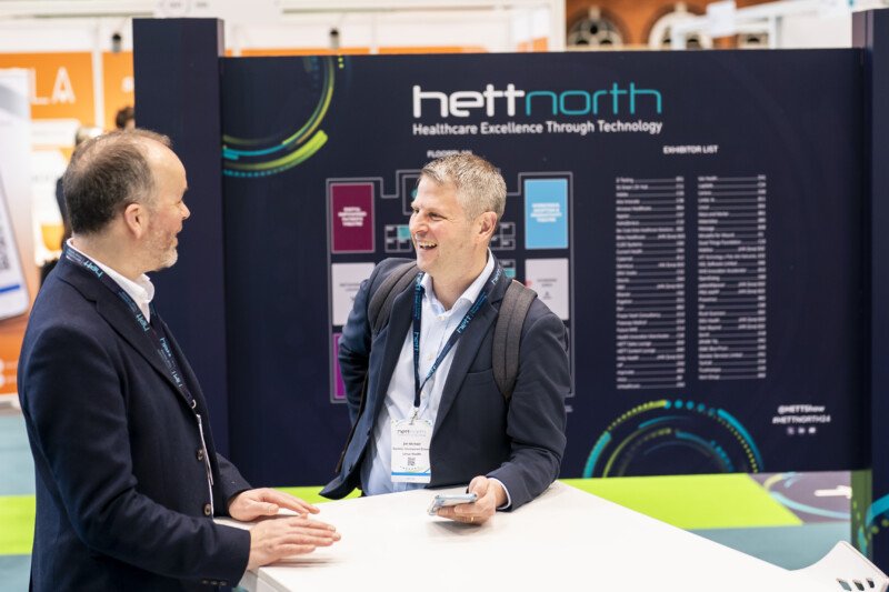 Two men, dressed in business attire, are engaged in a lively conversation at a booth with a sign reading "hett north" and "Healthcare Excellence Through Technology." Captured by a London conference photographer, one man holds a smartphone as both smile warmly