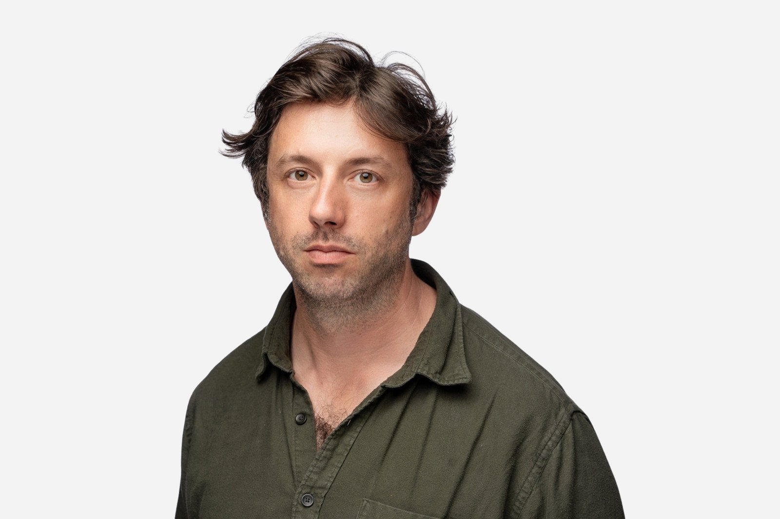 A man with wavy hair and a short beard is wearing a green button-up shirt. He is looking directly at the camera against a plain white background