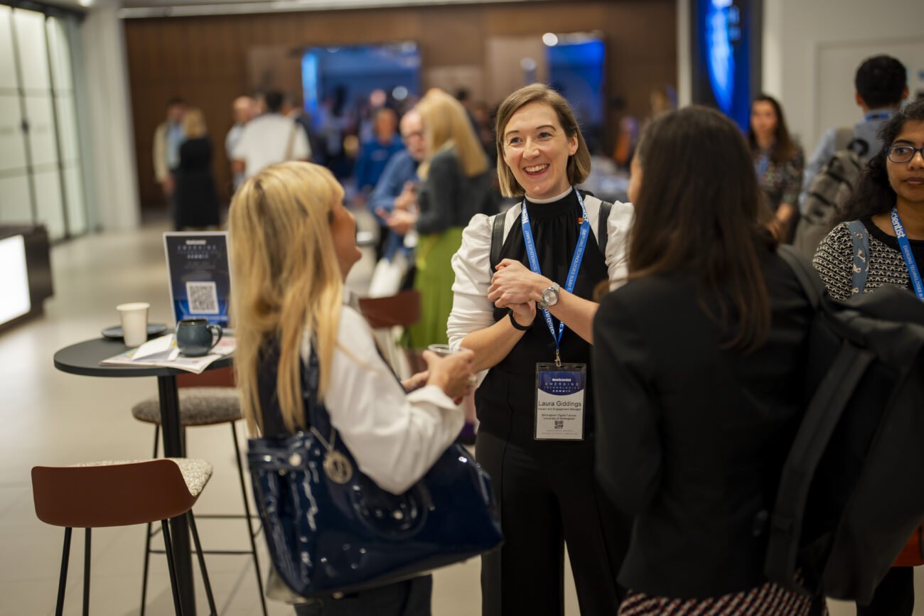 At a bustling London conference, a group of attendees is socializing. A woman with a name tag smiles warmly, shaking hands as the conference photographer captures the moment. In the background, tables laden with materials buzz with lively conversations among engaged participants