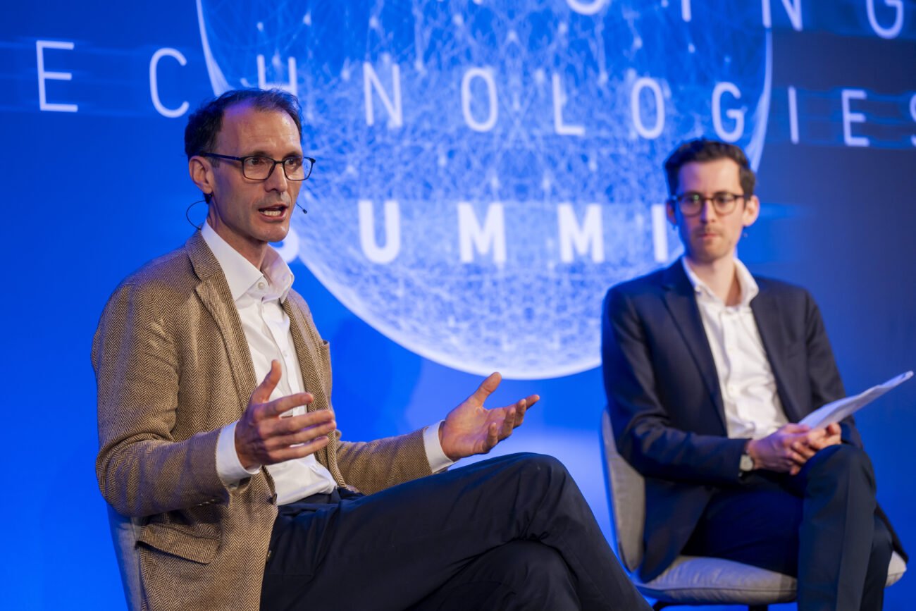 Two men in suits engage in discussion on stage at a London Technologies Summit. The man on the left gestures while speaking, and the man on the right holds a paper. A large, illuminated sign in the background reads "Technologies Summit," capturing a perfect moment for any conference photographer