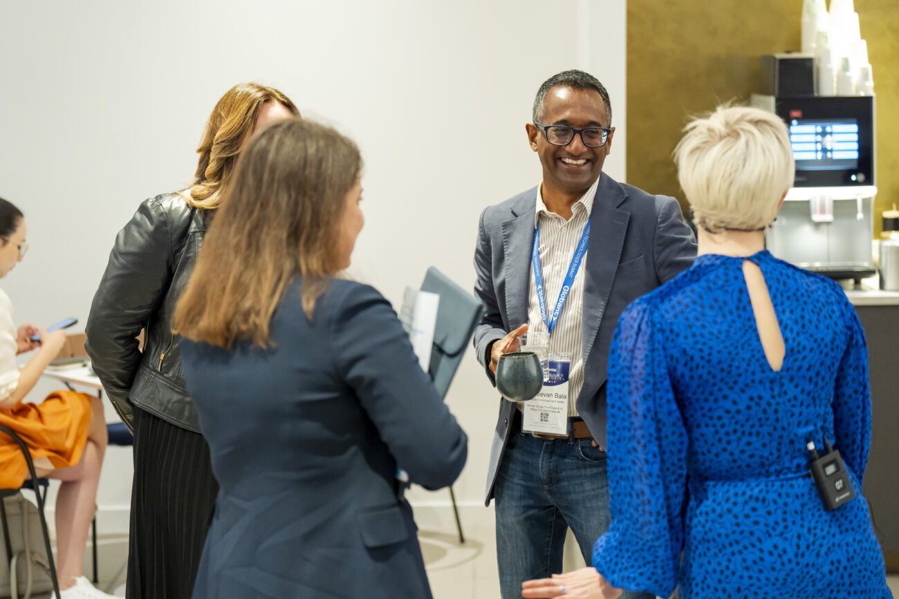 At a London conference, a photographer captures a lively office scene where a man with a lanyard and mug chats animatedly. The woman in the blue dress adds to the vibrant mix, while others casually converse, enhancing the lively atmosphere of collaboration