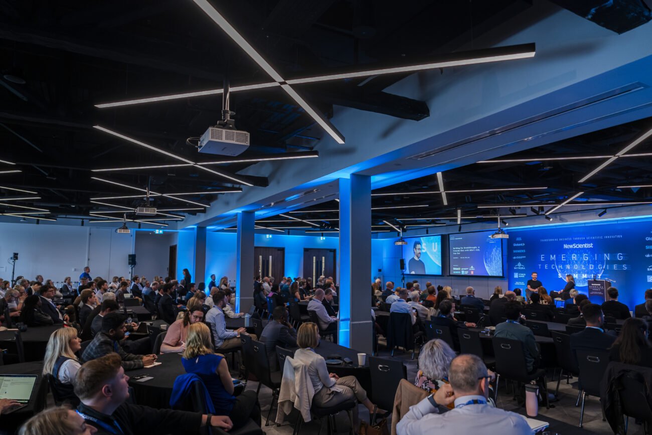 A large conference room brims with seated attendees, attentively listening to a panel discussion. Bathed in blue lighting, the room showcases presentations on vibrant screens. Captured by a London Conference Photographer, the engaged audience faces the stage where dynamic speakers hold sway