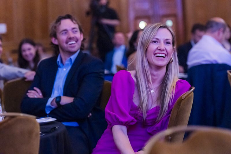 At a lively gathering, a woman in a purple dress and a man in a suit sit together, smiling and laughing. Captured by an event photographer, the scene showcases the vibrant essence of London corporate photography. James Gifford-Mead Photography - Event Photographer London
