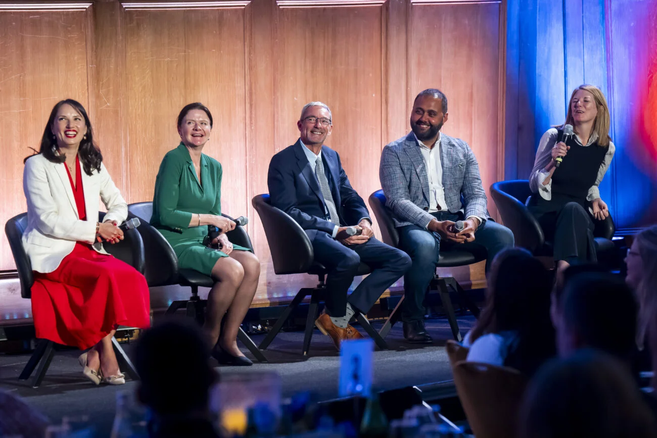 Five people sit on stage in a lively discussion panel, their smiles capturing the essence of corporate photography. One holds a microphone as warm lighting highlights the wooden paneling behind. Captured by an expert London event photographer, this scene offers an inviting glimpse into the event