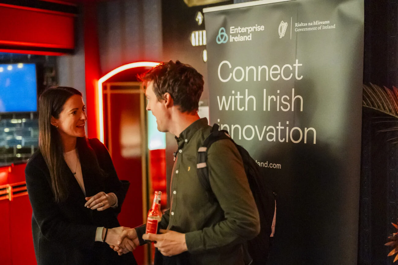 A man and woman shake hands at an event in front of a "Connect with Irish Innovation" banner, captured perfectly by a London Event Photographer. The man holds a drink, both in business attire amidst red and black decor, creating a business casual ambiance
