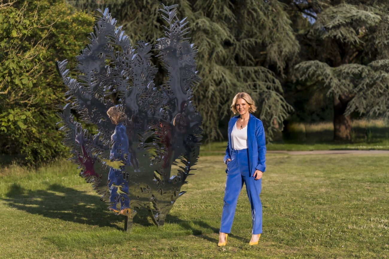 A corporate headshot photographer captures a person in a blue suit standing on grass beside a large abstract metallic sculpture, with tall trees forming a leafy backdrop under the sunny, clear sky. James Gifford-Mead Photography - Event Photographer London