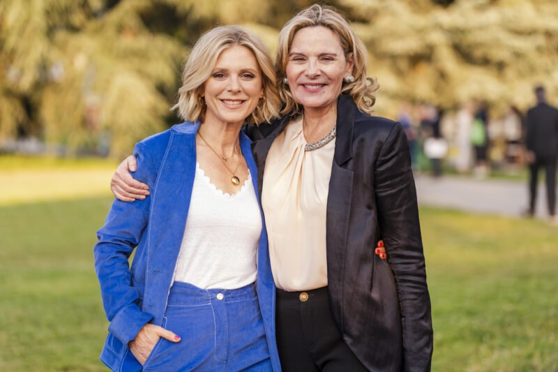 Two women, captured by a skilled event photographer in London, stand closely in a park, smiling at the camera. One wears a blue suit over a white top, and the other dons a black blazer over a cream blouse. They look relaxed and friendly, with blurred greenery providing the backdrop. James Gifford-Mead Photography - Event Photographer London