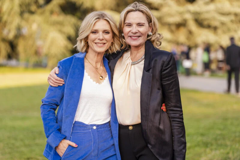 Two women, captured by a skilled event photographer in London, stand closely in a park, smiling at the camera. One wears a blue suit over a white top, and the other dons a black blazer over a cream blouse. They look relaxed and friendly, with blurred greenery providing the backdrop