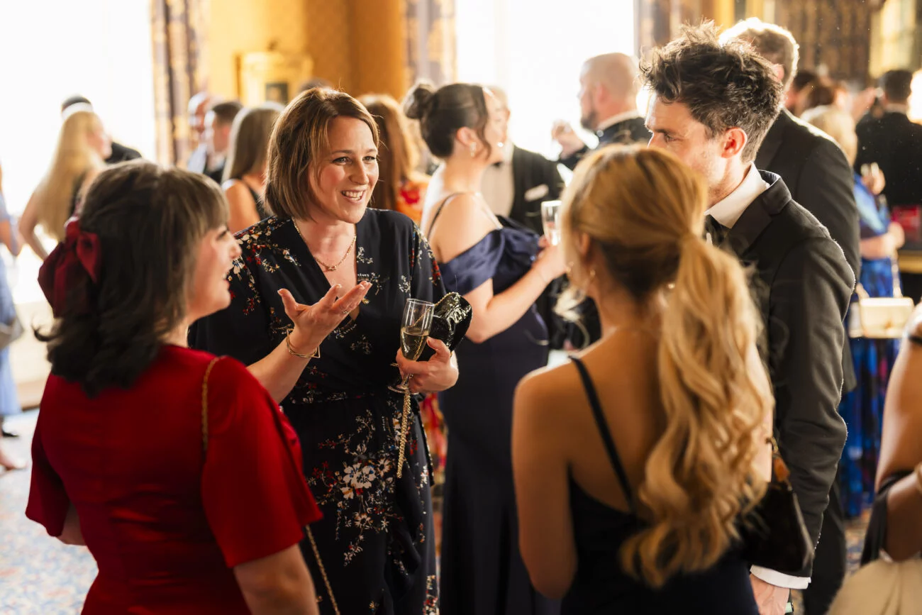 Amid the elegant setting of a formal event, well-dressed individuals are captured by an expert London event photographer. One woman, dressed in a floral gown and holding a wine glass, animates the lively conversation, all beautifully framed in soft lighting and decorative surroundings
