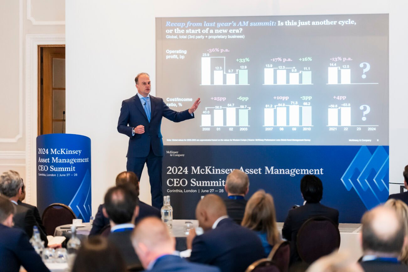 A speaker in a suit stands beside a chart on screen, presenting at the 2024 McKinsey Asset Management CEO Summit in London. Attendees sit at round tables, viewing the presentation, all captured by an expert London event photographer specializing in corporate photography. James Gifford-Mead Photography - Event Photographer London