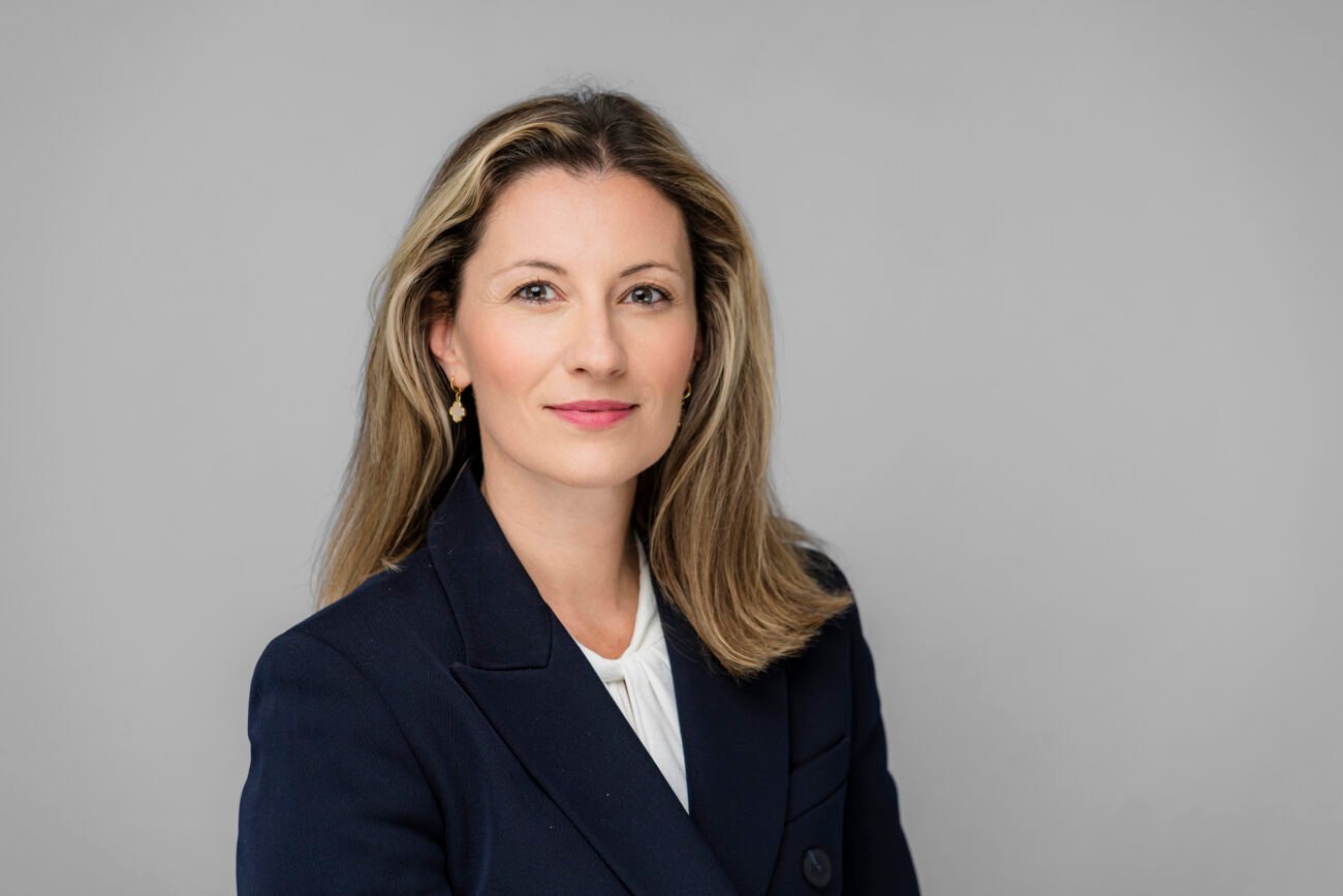 A woman with long, light brown hair poses against a gray background, embodying the essence of corporate photography. She wears a dark blazer over a white blouse and small earrings, her expression neutral. James Gifford-Mead Photography - Event Photographer London