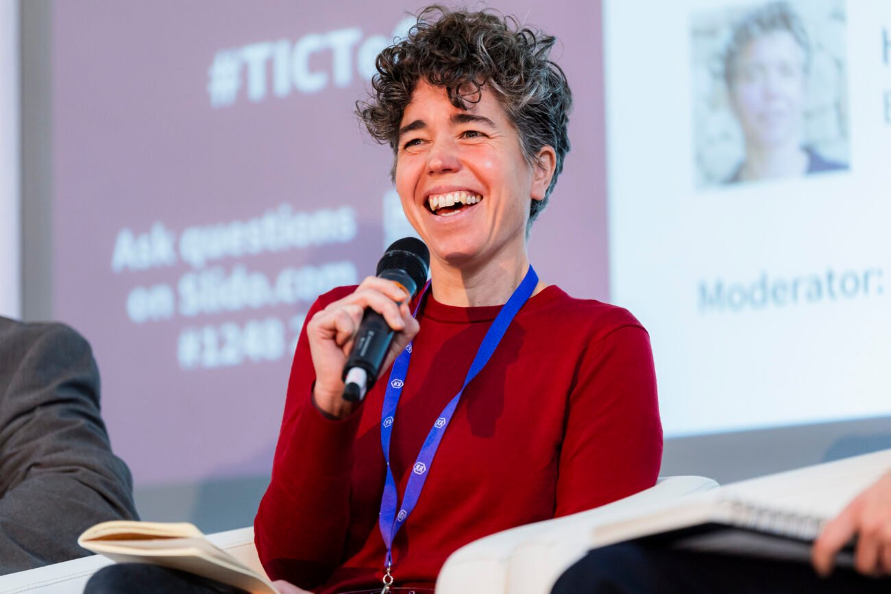 A person with curly hair, wearing a red sweater and blue lanyard, is smiling while holding a microphone. They're captured brilliantly by a London Event Photographer in an event setting, with a blurred presentation screen in the background.