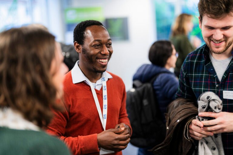 A group of people are engaged in conversation at an indoor event. A man in a red sweater smiles, wearing a name tag. People around him are also smiling and chatting. The setting appears casual and social, with blurred backgrounds