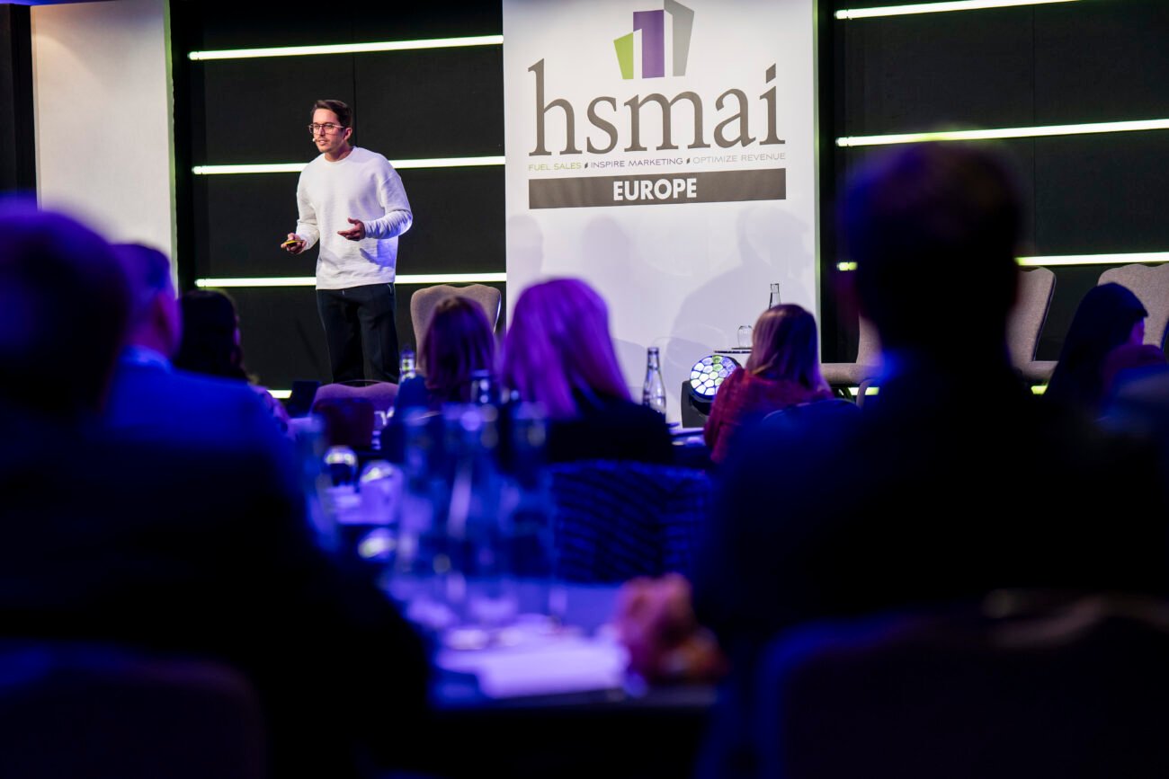 A speaker in a white sweater presents at an HSMAI Europe conference. The audience is seated, facing him, with focus on a large banner featuring the HSMAI logo. The room is dimly lit, creating a formal and engaged atmosphere.