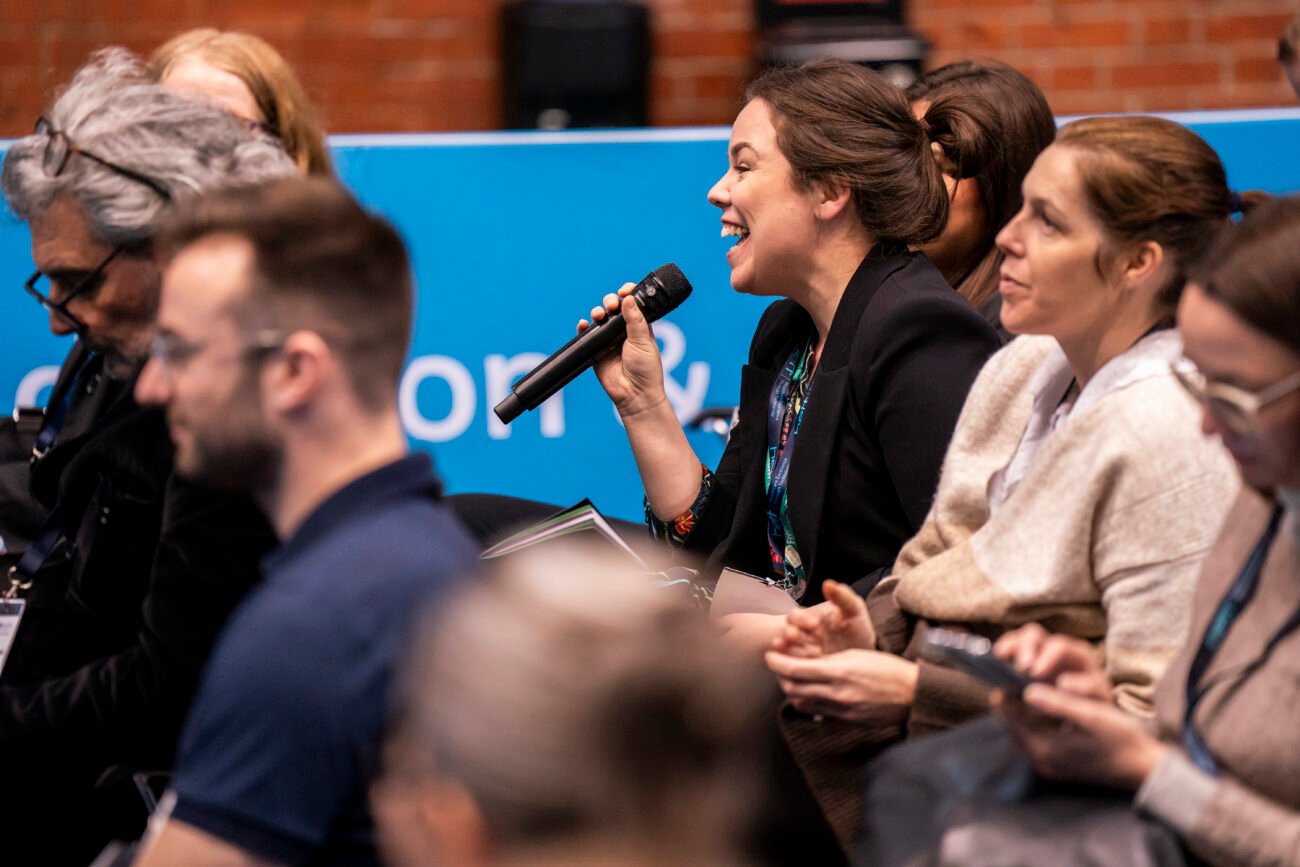 A woman in a black blazer, mid-discussion, holds a microphone among other seated attendees at a conference. She appears engaged while others listen or use devices. A blue banner with unreadable text is in the background