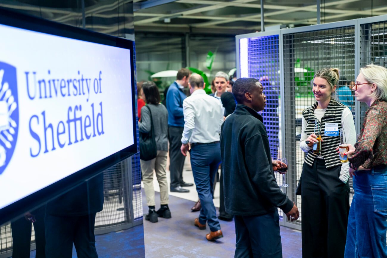 A group of people socializing at an event, with a screen displaying "University of Sheffield" on the left. The setting is modern, with metal mesh panels and overhead lights. Some individuals hold drinks as they engage in conversation
