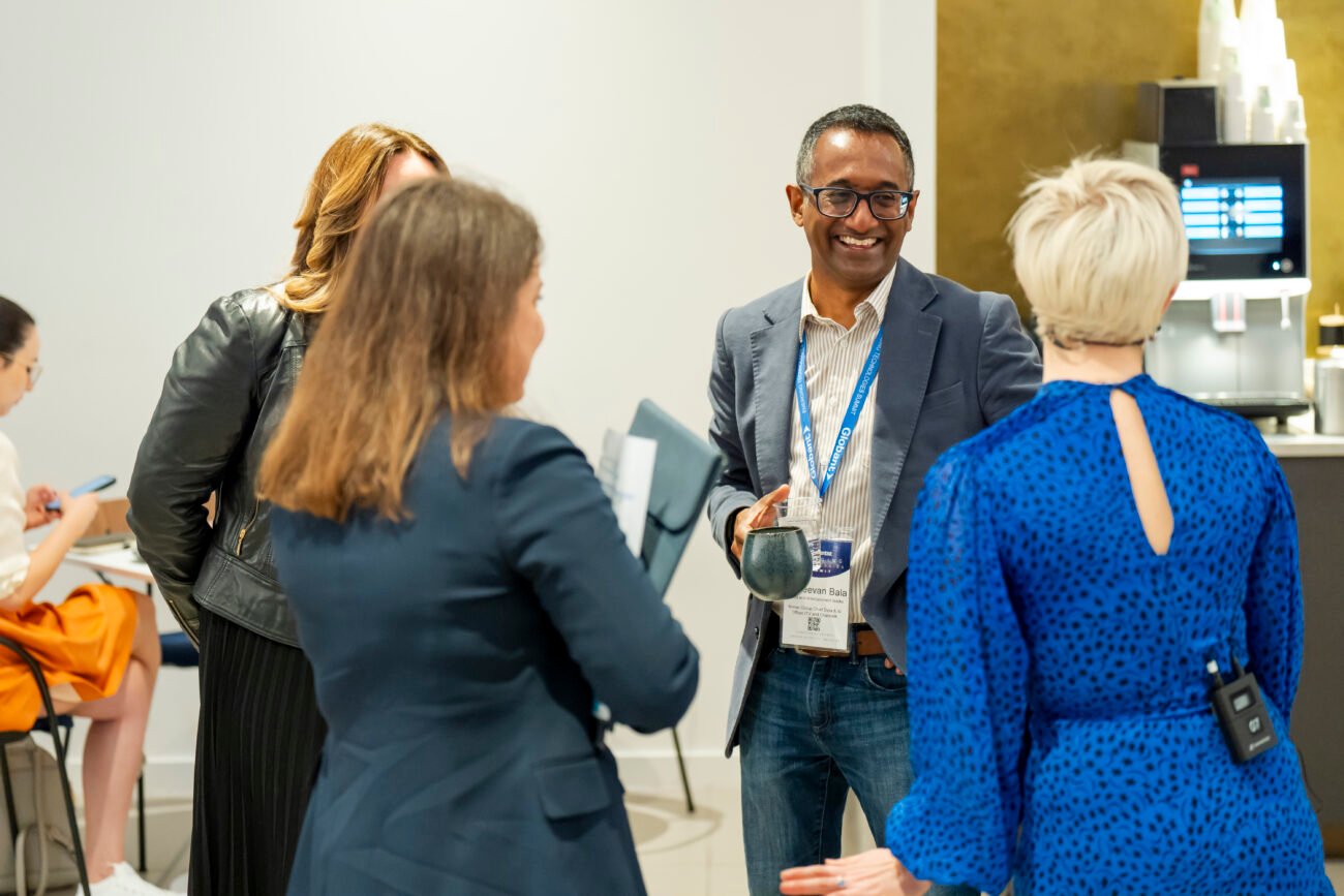 A group of four people in business attire engage in a friendly conversation, smiling and holding coffee cups. A person with short hair in a blue dress is in the foreground, while a person holds a tablet in the background. James Gifford-Mead Photography - Event Photographer London