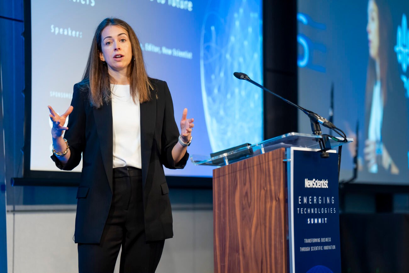 A woman stands at a podium, speaking into a microphone. She is gesturing with both hands. Behind her is a large screen displaying a blue and white graphic with text. The event is the Emerging Technologies Summit.