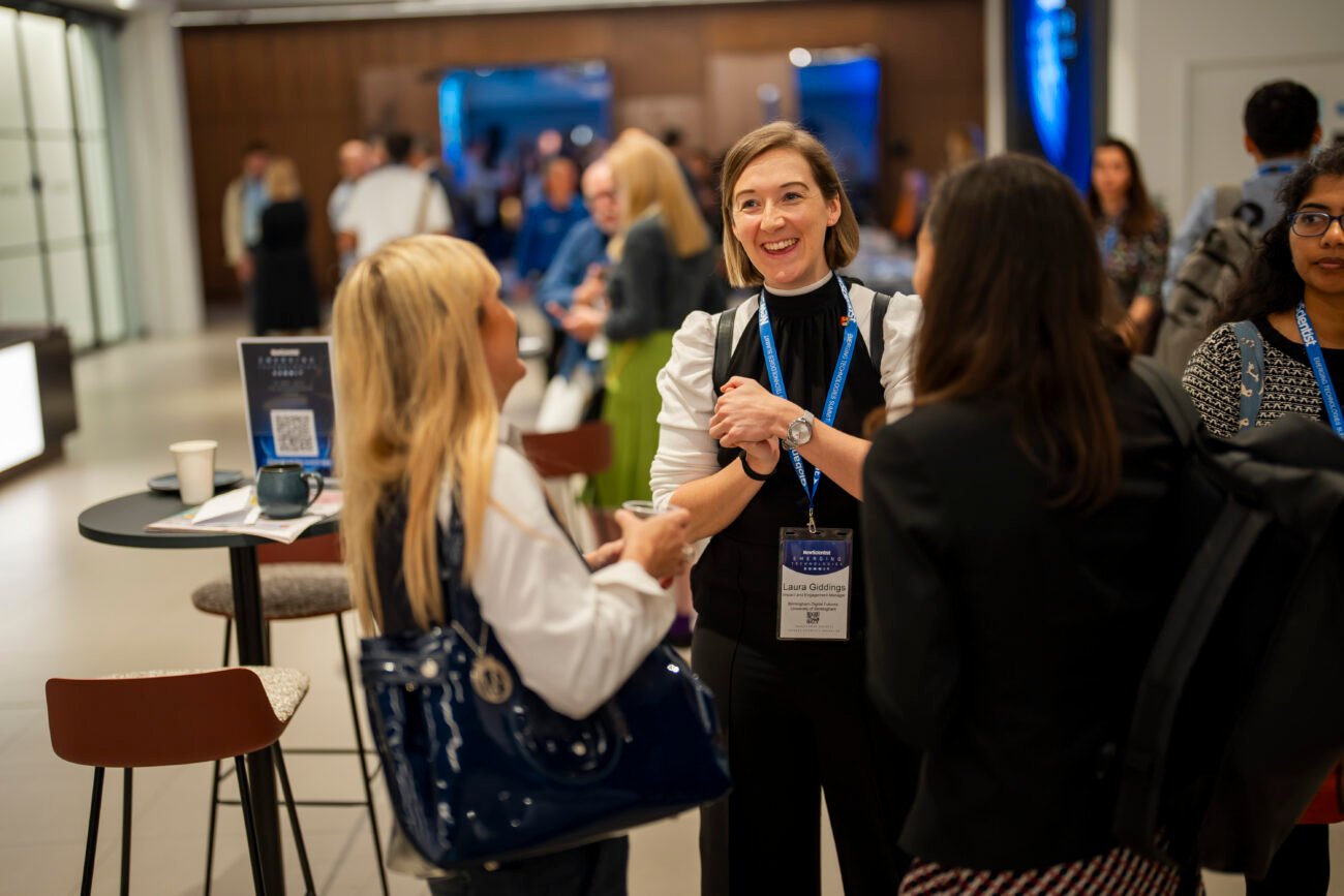 A group of people is engaged in conversation at a networking event. They are standing indoors, wearing business attire and event badges. A table with a coffee cup and a brochure is nearby. The atmosphere is lively and professional. James Gifford-Mead Photography - Event Photographer London