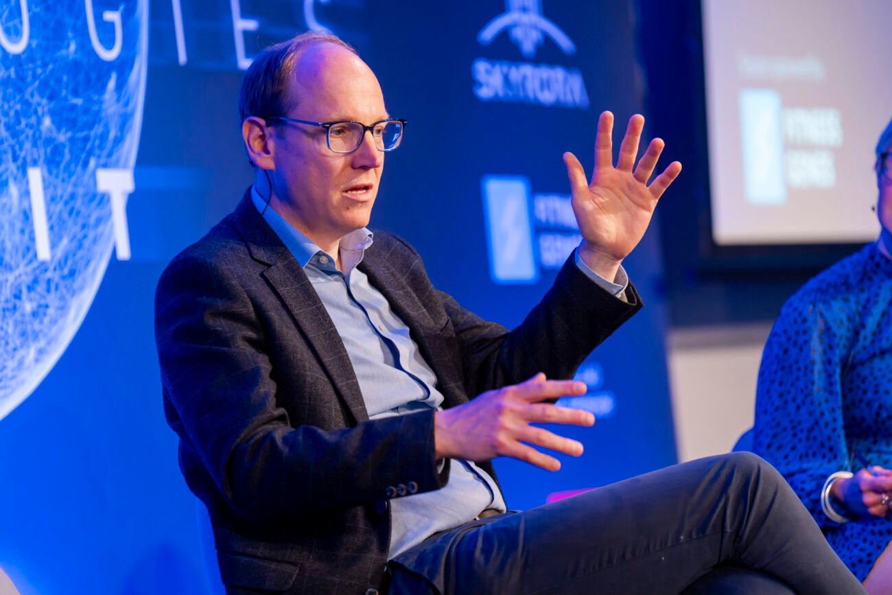 A person with glasses speaks animatedly, gesturing with both hands, while seated on a stage. They are wearing a suit jacket over a light blue shirt. The background has a blue hue with partial text and logos visible. James Gifford-Mead Photography - Event Photographer London